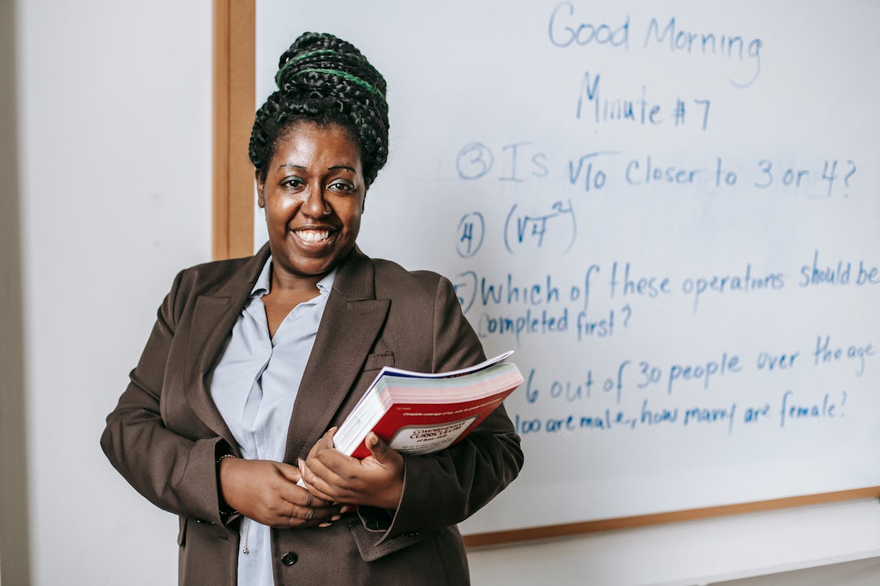 A smiling teacher holding textbooks stands confidently in front of a whiteboard.